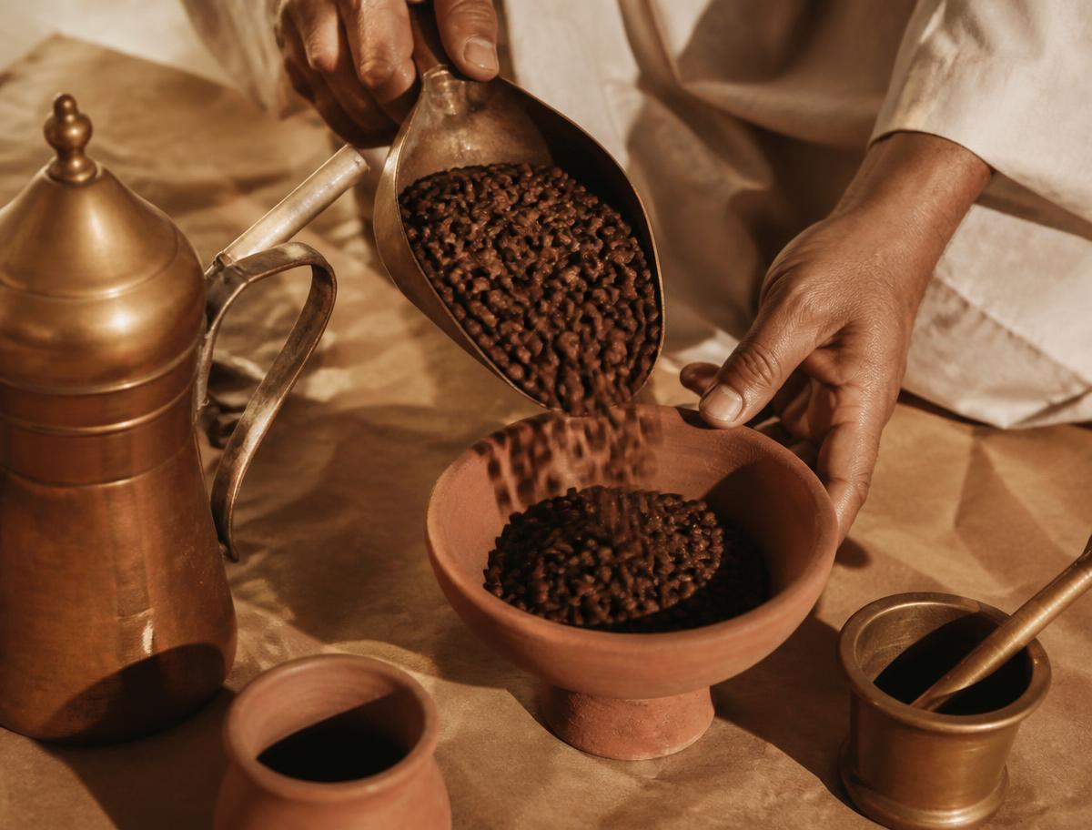 يد تصب بن محمص في وعاء فخاري بجانب أدوات قهوة نحاسية